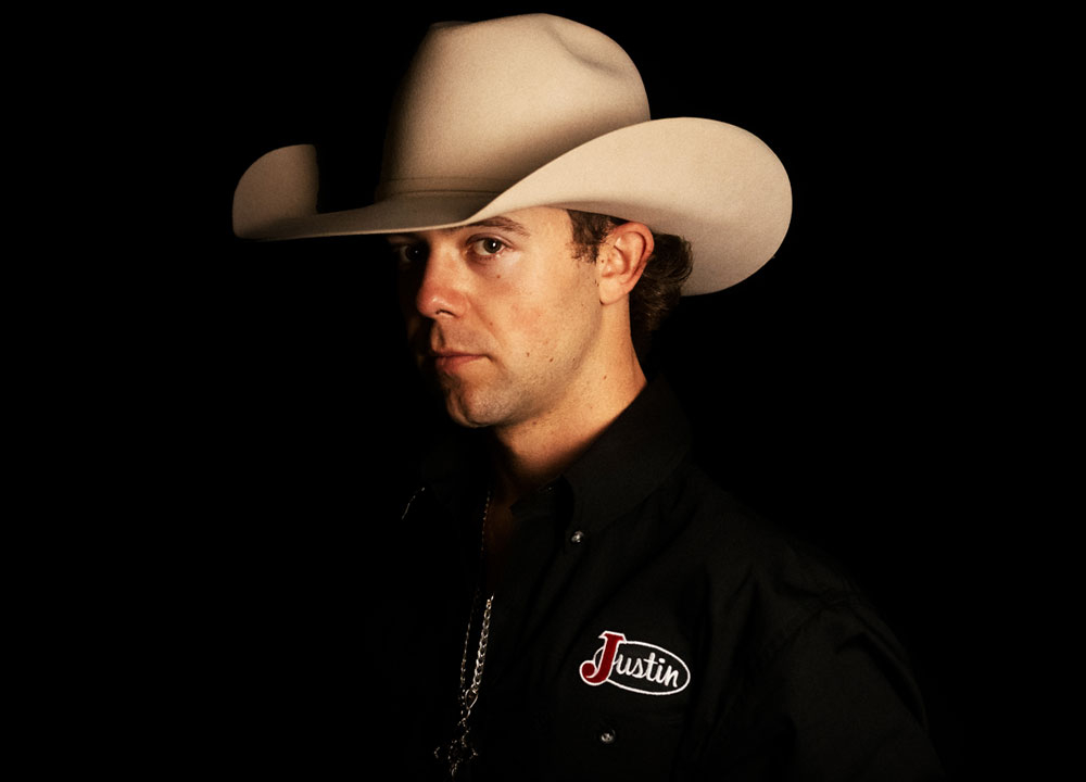 Hayes Weight poses in a studio portrait wearing a white cowboy hat and Justin-branded shirt against a dark background.