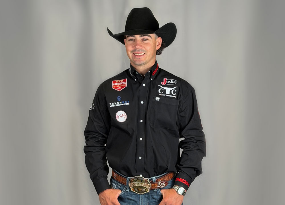 Damian Brennan poses in a portrait wearing a black cowboy hat and sponsor-branded rodeo shirt with a championship belt buckle against a neutral backdrop.

