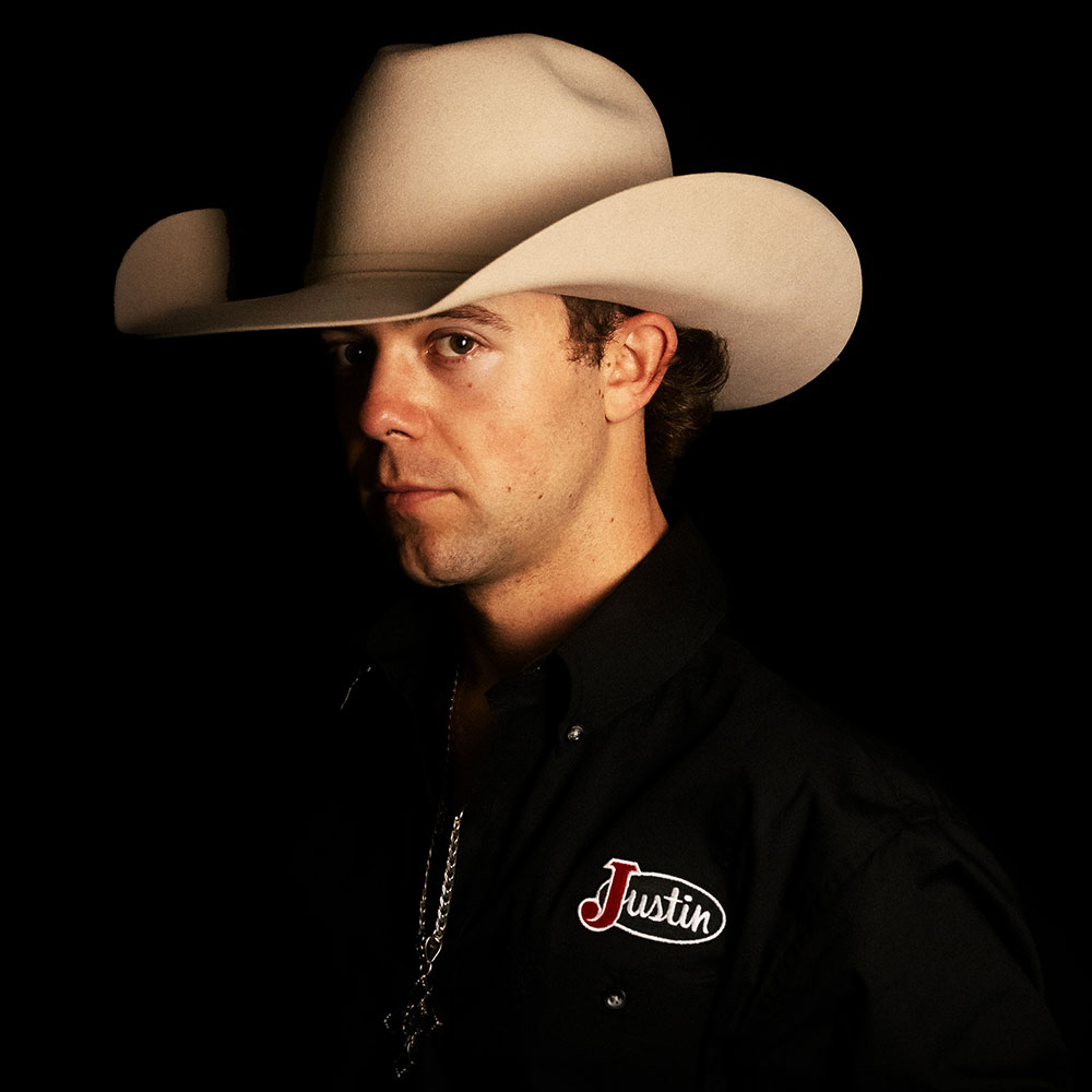 Hayes Weight poses in a studio portrait wearing a white cowboy hat and Justin-branded shirt against a dark background.
