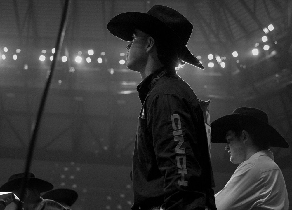 Damian Brennan stands in profile beneath arena lights at the Fort Worth Stock Show & Rodeo, wearing a black cowboy hat and rodeo gear while waiting near the chutes before competition.
