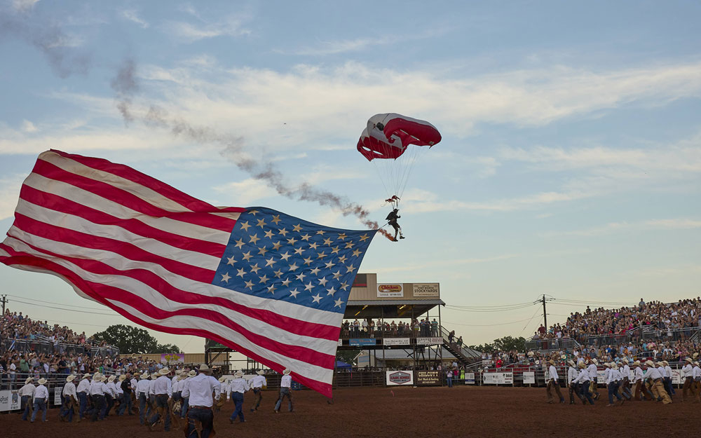 A skydiver descends into a packed rodeo arena at sunset, trailing a massive American flag and red smoke.