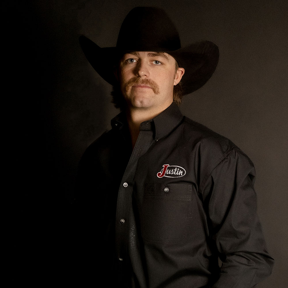 Wyatt Casper poses in a studio portrait wearing a black cowboy hat and Justin-branded shirt with a championship belt buckle against a dark background.