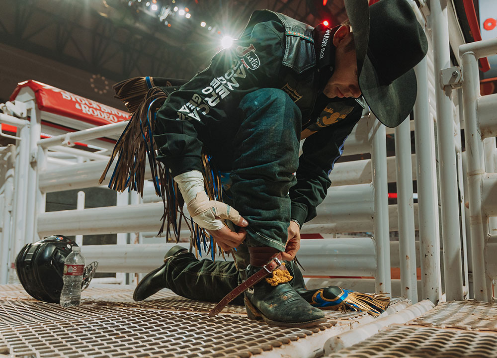 Hayes Weight kneels near the chutes at a rodeo arena, tightening his boot strap while wearing a cowboy hat and rodeo gear under bright arena lights.