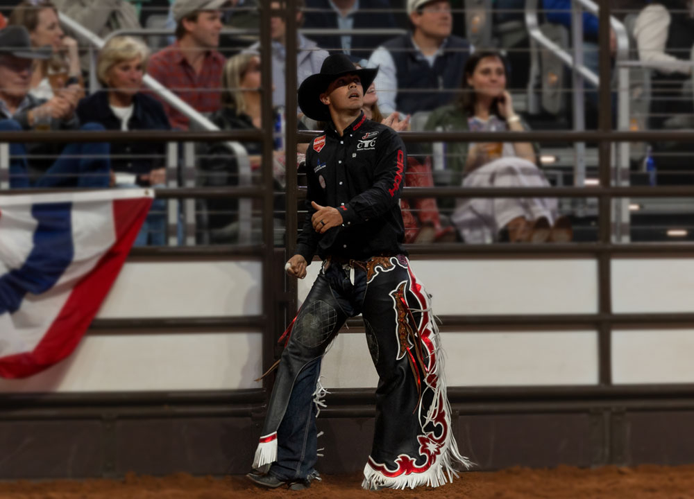 Damian Brennan stands in the arena at the Fort Worth Stock Show & Rodeo wearing a black cowboy hat and chaps, looking toward the replay screen after riding in the saddle bronc event.