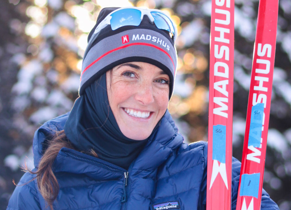 Skier smiling while holding red Madshus skis in a snowy forest setting, wearing a blue insulated jacket, winter hat, and sunglasses, with sunlight filtering through the trees in the background.