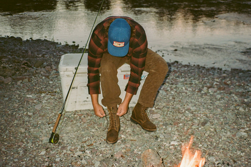 A man wearing The Trailboss boots in brown sitting on a cooler by a river lacing up his boots.