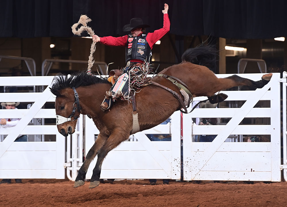 Wyatt Casper compite en saddle bronc, montando un caballo bronco en la arena mientras mantiene su mano libre en alto durante la monta.