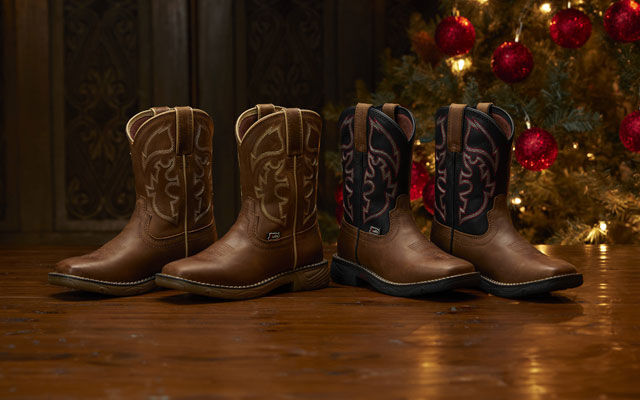 Two pairs of kids boots sitting on a wood floor in front of a Christmas tree.