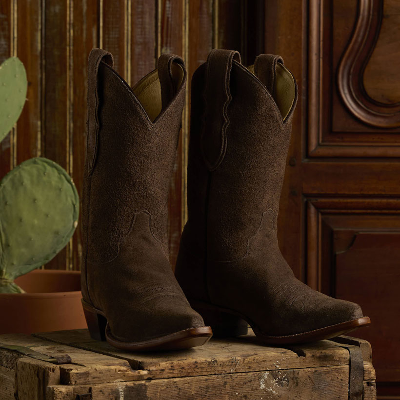The Reba By Justin Fancy 11" Suede boots in Mocha Brown sitting on a table next to a cactus.