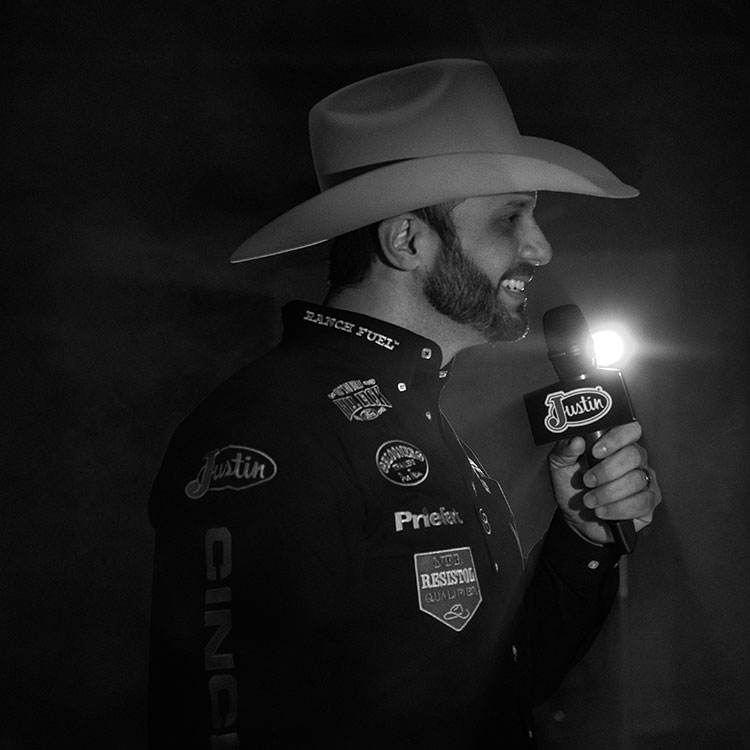 Anthony Lucia wearing a black shirt and cowboy hat, holding a microphone and talking with a light behind him. 