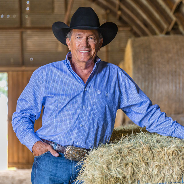 A man, George Strait wearing a cowboy hat while standing next to a bale of hay in a barn.