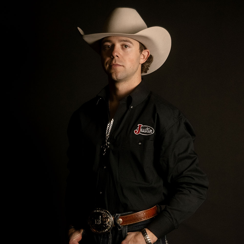 Hayes Weight poses in a studio portrait wearing a white cowboy hat and Justin-branded shirt against a dark background.