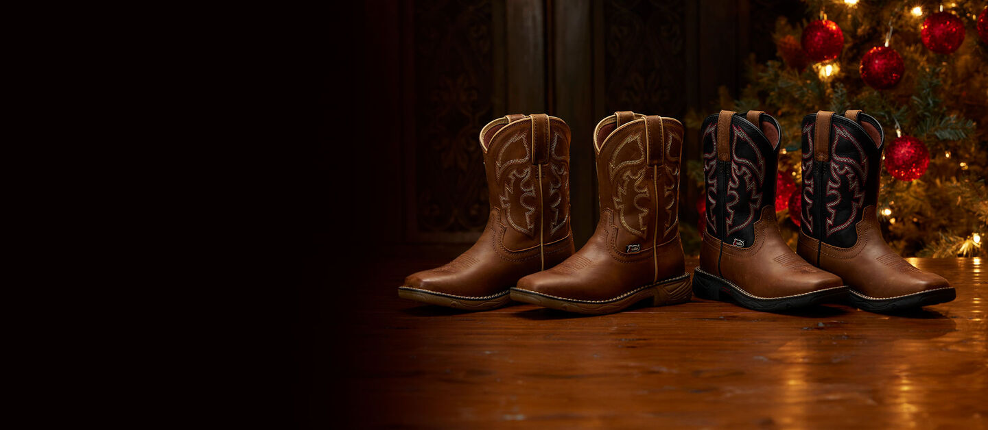 Two pairs of kids boots sitting on a wood floor in front of a Christmas tree.