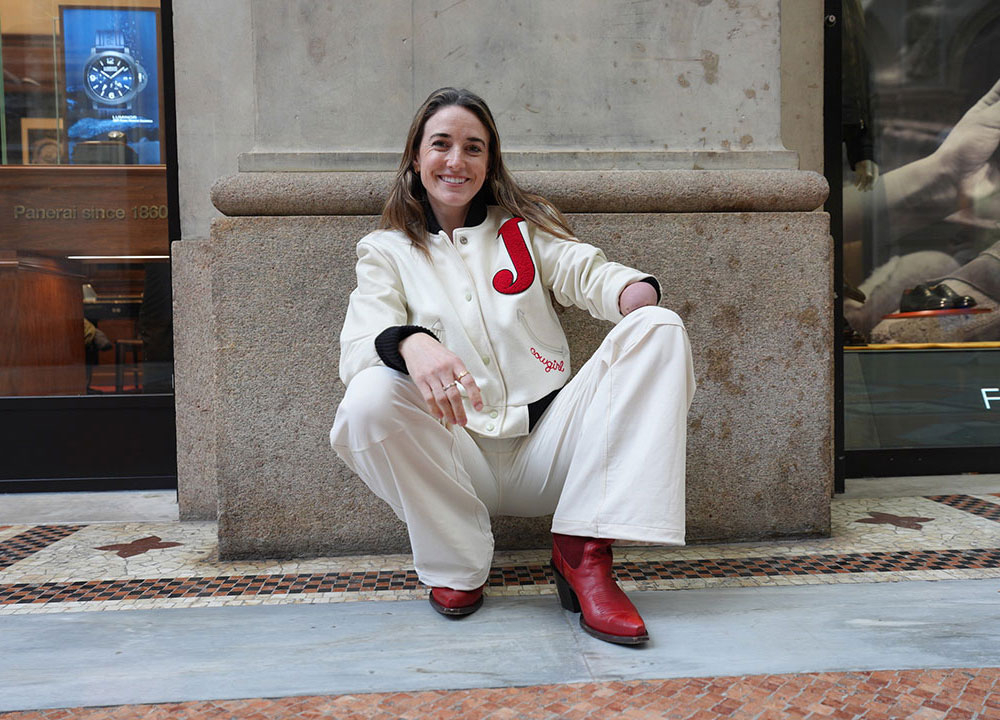 Smiling woman sitting on a stone ledge in an indoor shopping area, wearing a cream jacket with a red letter patch, matching pants, and red heeled boots, with storefront displays visible in the background.