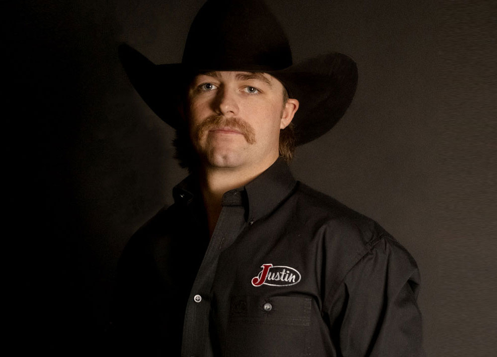Wyatt Casper posa en un retrato de estudio vistiendo un sombrero vaquero negro y una camisa con la marca Justin, con una hebilla de campeonato, sobre un fondo oscuro.