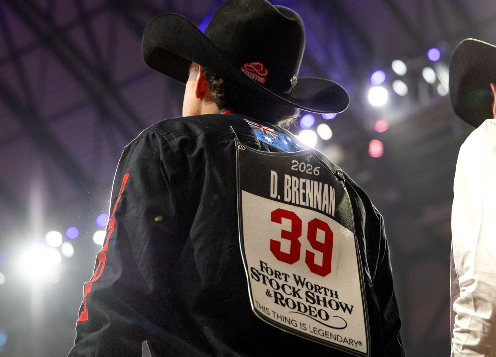 Damian Brennan stands near the chutes at the Fort Worth Stock Show & Rodeo wearing a black cowboy hat and competition vest with his name and number displayed under the arena lights.
