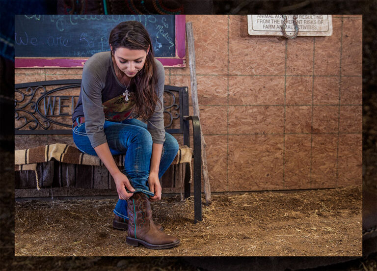 A woman sitting on a bench in a barn putting on Justin Gypsy boots.
