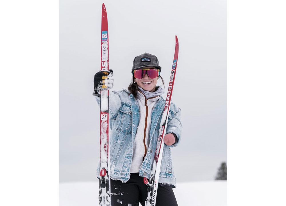 Smiling skier standing in a snowy landscape, holding red Madshus skis while wearing a denim jacket, winter layers, a black cap, and reflective snow goggles.