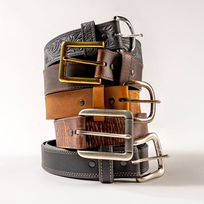 The Scroll Tooled Belt in black, Sycamore Cinch Belt in brown, Work Basic Belt in tan, Bomber Belt in brown, and Carson Belt in black stacked in a pile.