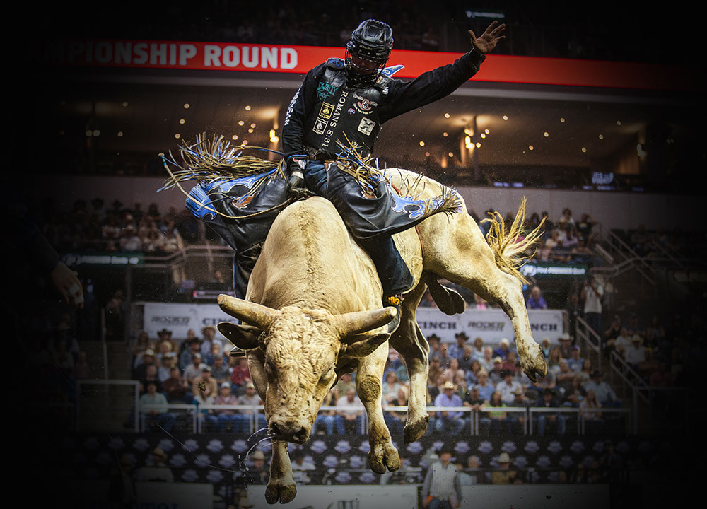 Hayes Weight competes in bull riding during a championship round, riding a bucking bull in front of a packed arena crowd under bright rodeo lights.