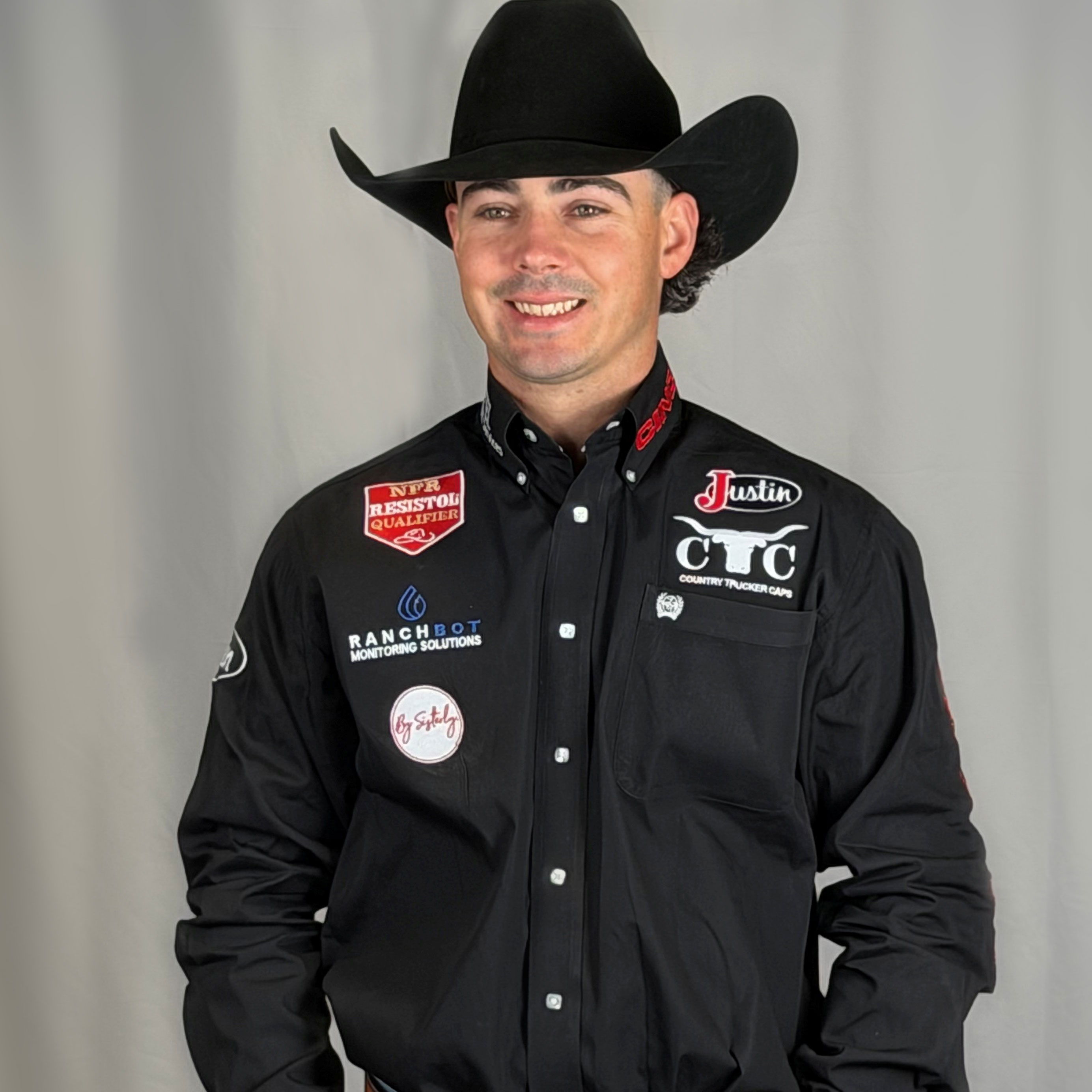 Damian Brennan poses in a portrait wearing a black cowboy hat and sponsor-branded rodeo shirt with a championship belt buckle against a neutral backdrop.