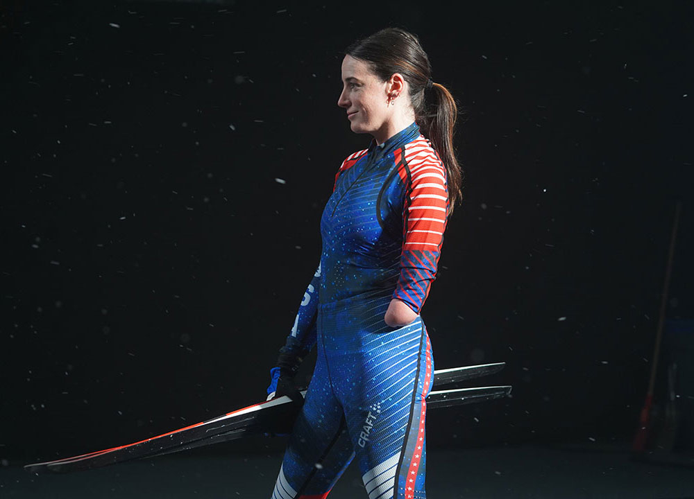 Side-profile portrait of a skier wearing a blue and red racing suit, holding skis against a dark background with light snow falling around her.