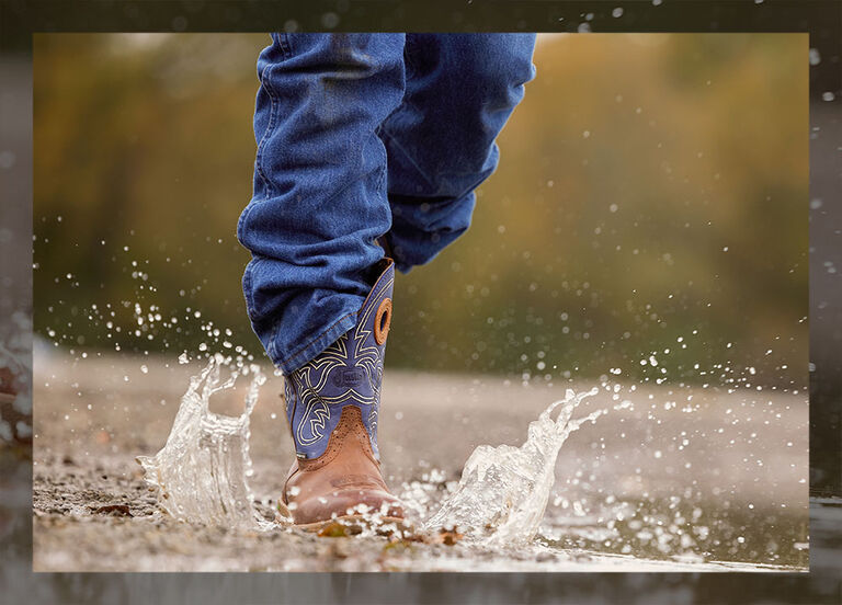 A blue and brown waterproof Justin Nitread boot being worn by a model and walking through a puddle of water.