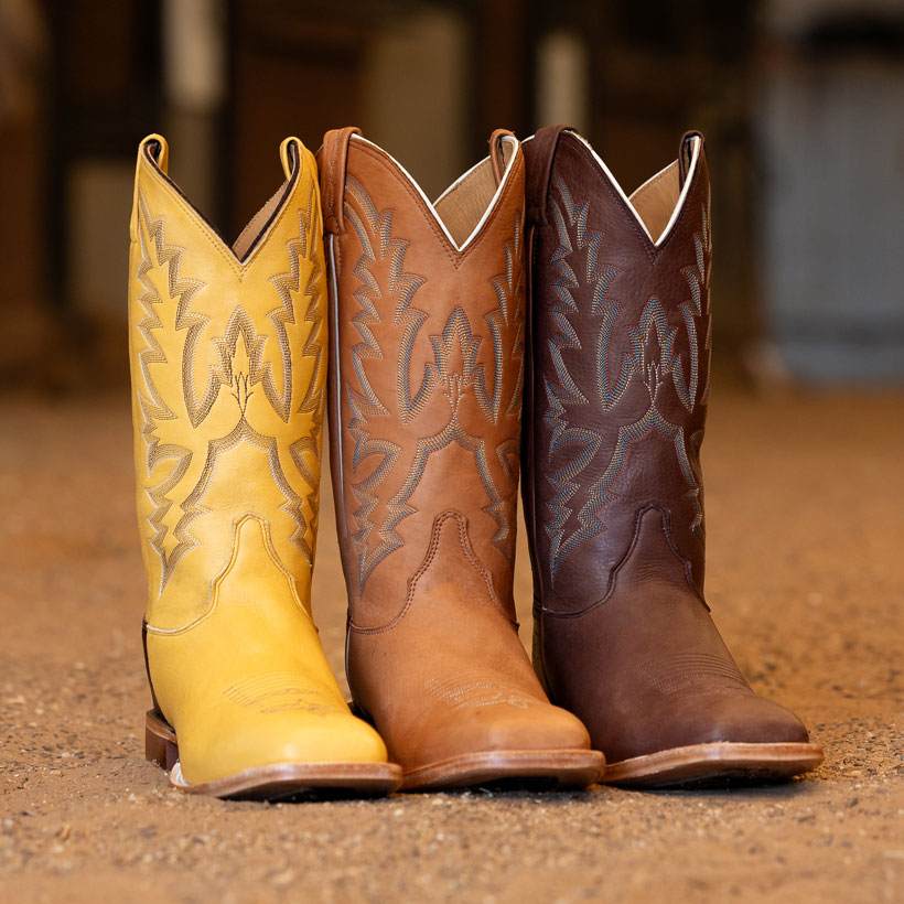 Botas vaqueras Boston de 13 pulgadas, modelo Whitetail Deer, en colores chocolate (marr&oacute;n), amarillo y marr&oacute;n co&ntilde;ac, sobre un suelo de tierra.