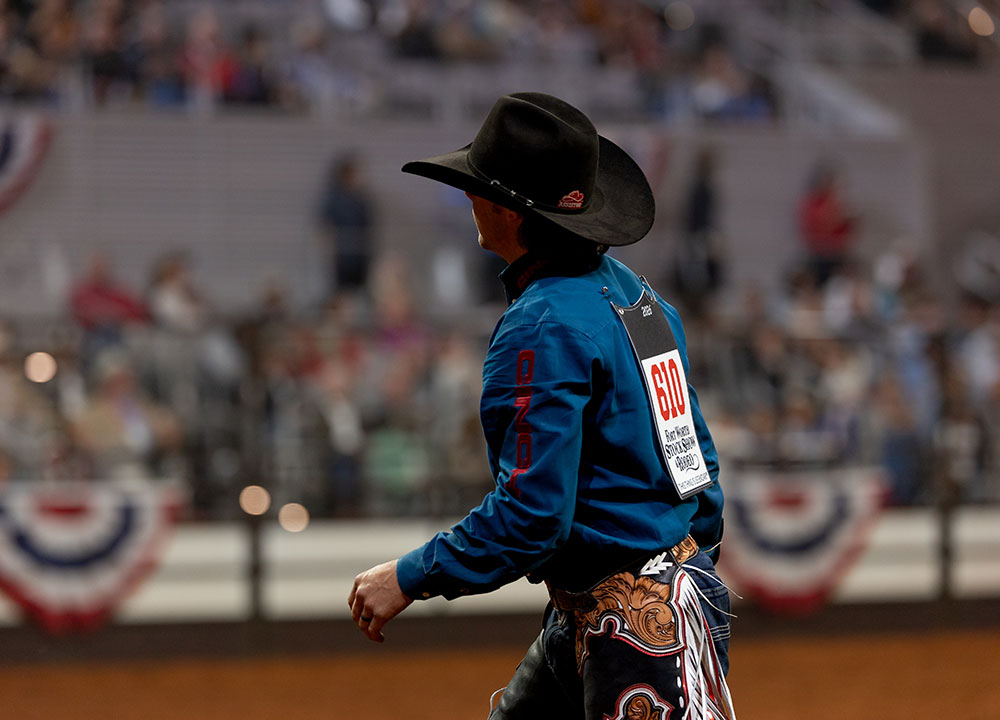 Wyatt Casper camina por la arena del Fort Worth Stock Show & Rodeo vistiendo una camisa azul de rodeo, sombrero vaquero negro y su n&uacute;mero de competencia en la espalda.