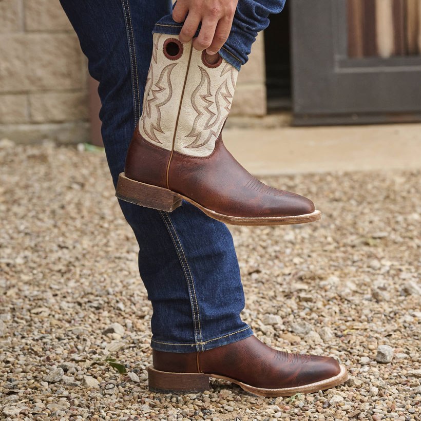 A man pulling on a Men’s Caddo 11” Western boot in cognac damiana brown while leaning against a tractor