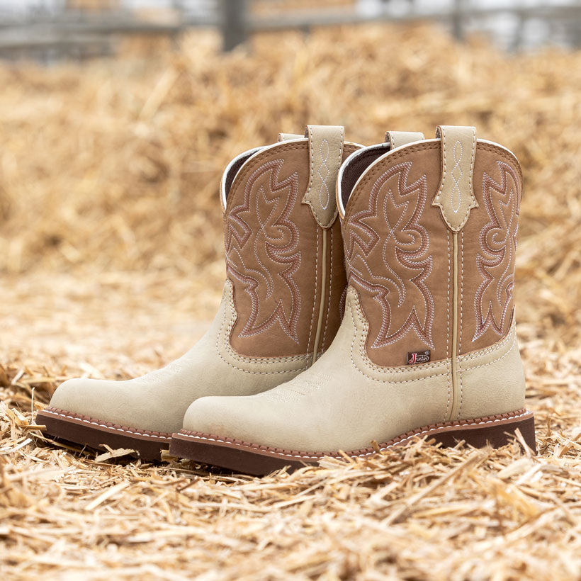 A pair of light tan and brown Gypsy Western boots with decorative stitching sit on a bed of straw in front of metal livestock pens.