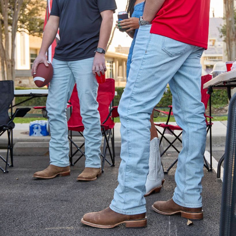 A group of people tailgating in a parking lot, holding drinks and a football, wearing Justin light wash jeans and brown Frontier boots. 