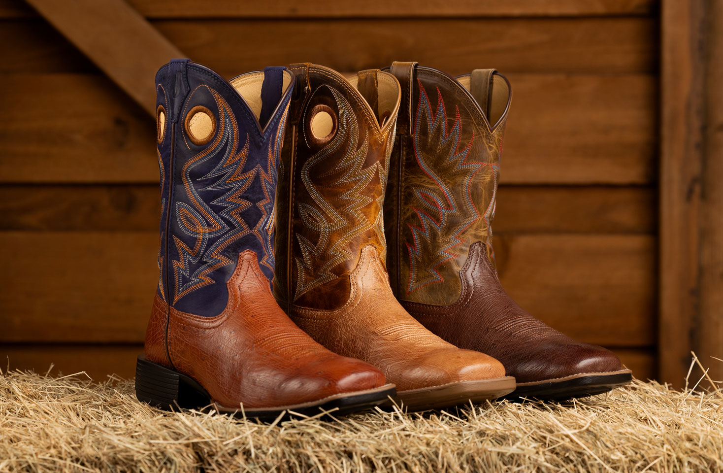 Men’s Conroe 11” smooth Ostrich in brown and Roswell 11” Smooth Ostrich in brown posed on a hay bale in front of a wooden background.