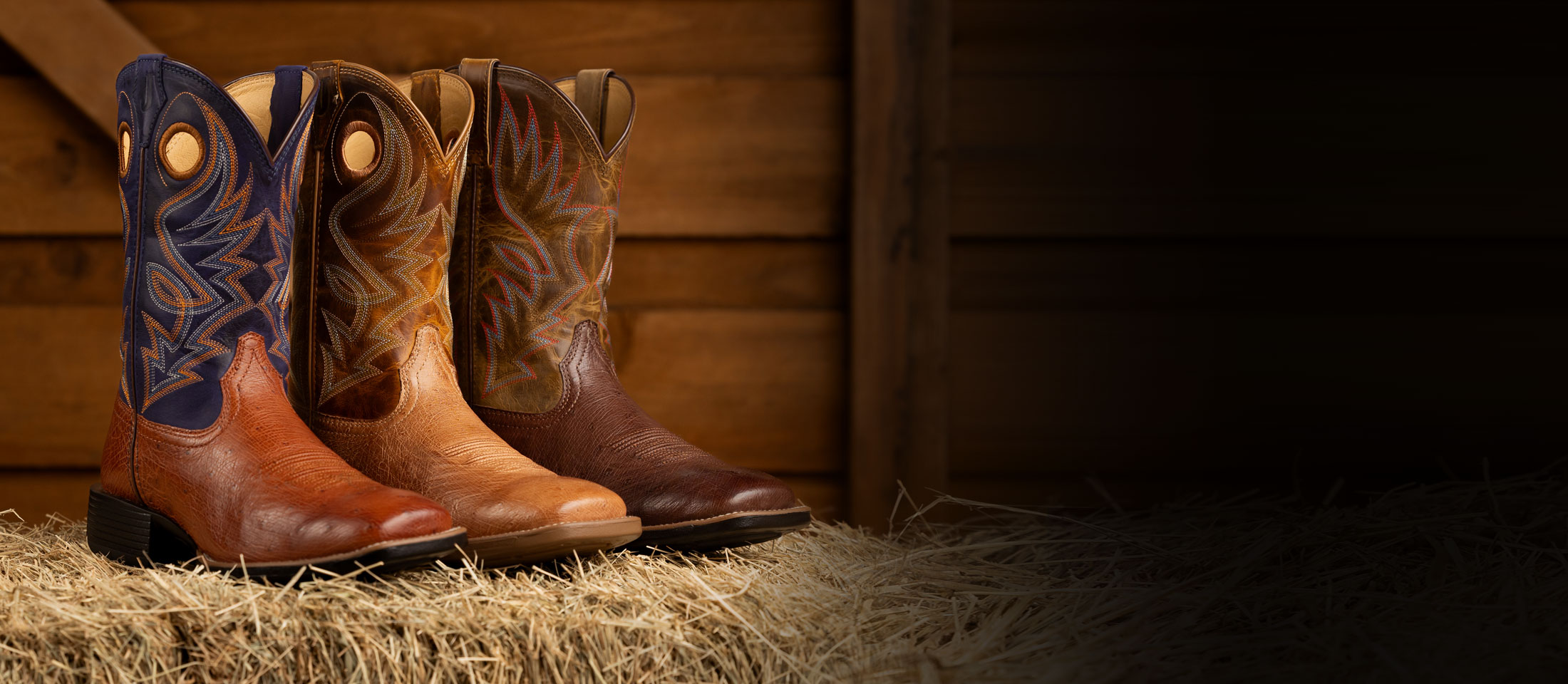 Men’s Conroe 11” smooth Ostrich in brown and Roswell 11” Smooth Ostrich in brown posed on a hay bale in front of a wooden background.