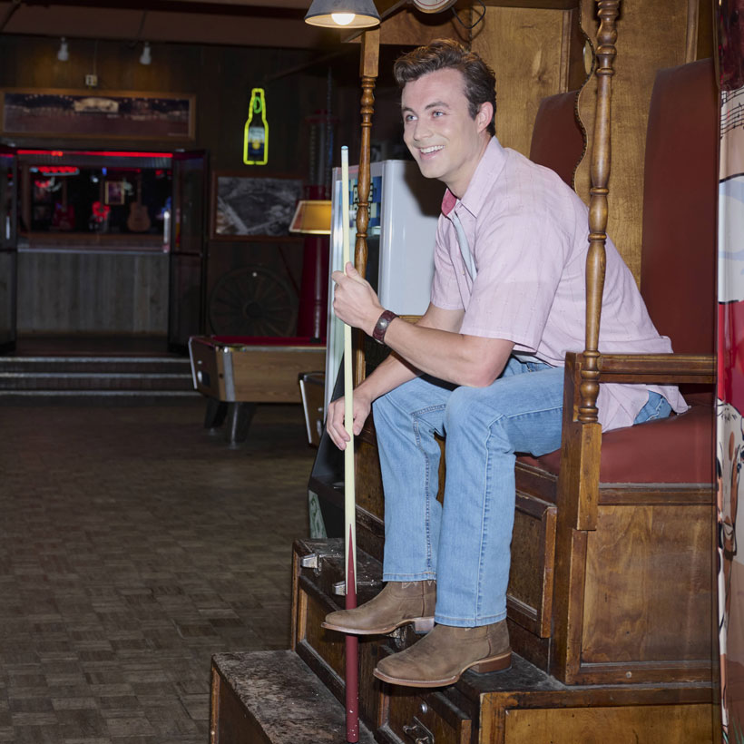 A smiling man in a pink shirt, jeans, and Frontier boots sits on a tall wooden chair holding a pool cue in a dim bar.