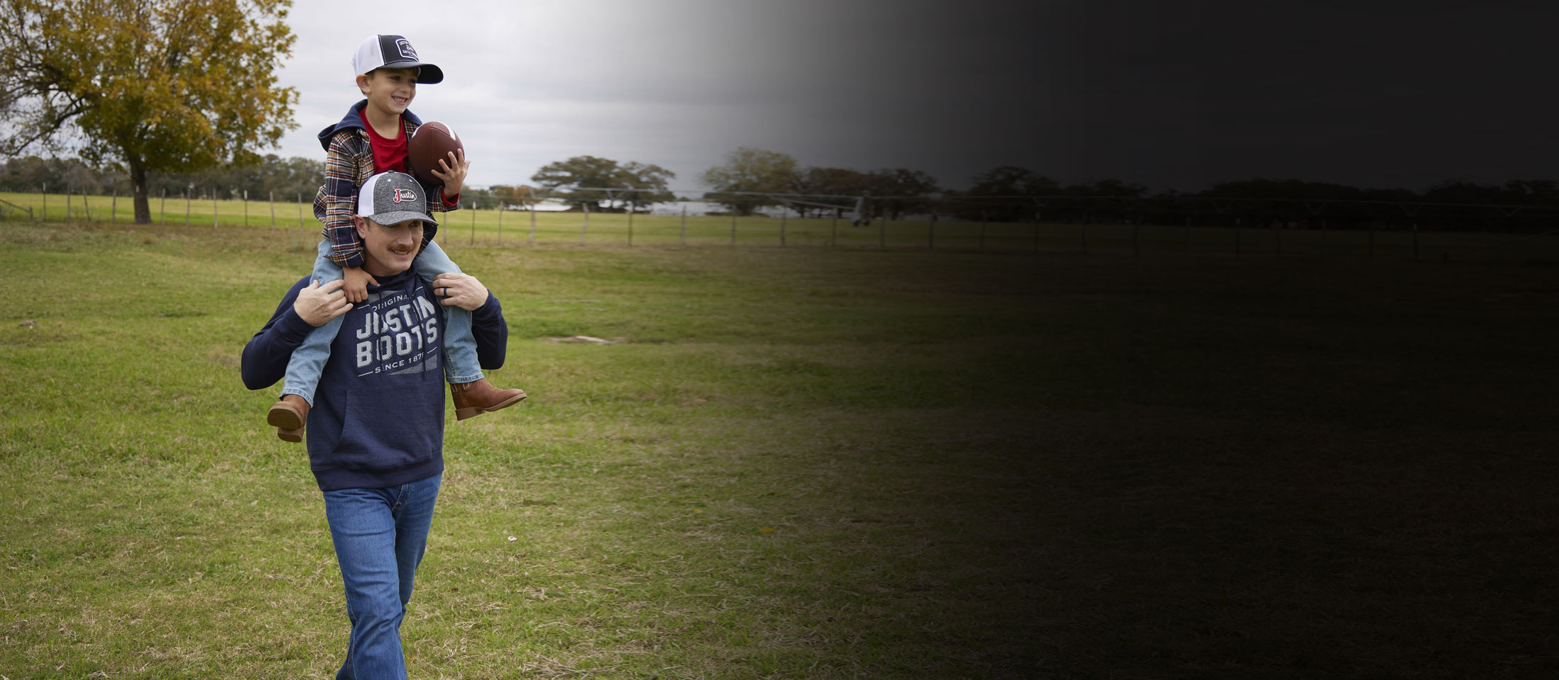 A father carrying his son who is holding a football, sitting on his dad’s shoulders, in a green field, laughing. 