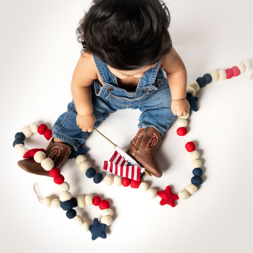 A baby with black hair is sitting wearing Baby Canter baby boots in brown and red, holding an American flag. 