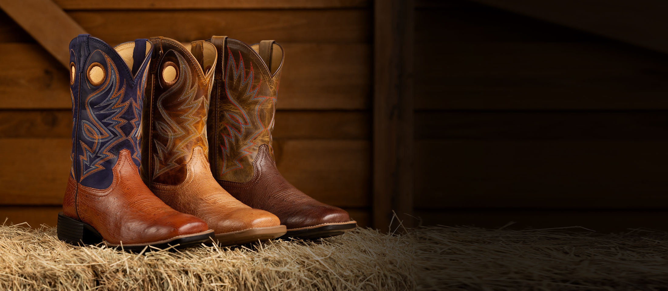 Men’s Conroe 11” smooth Ostrich in brown and Roswell 11” Smooth Ostrich in brown posed on a hay bale in front of a wooden background. 