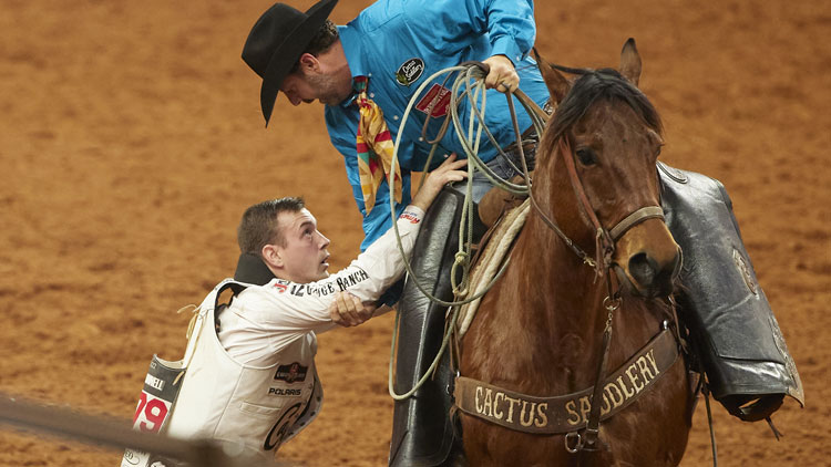 A pickup man is helping Tim O’Connell off his horse at the rodeo.