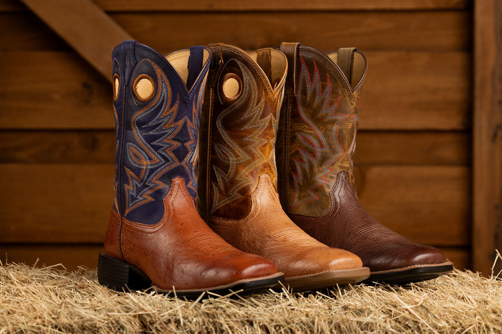 Men’s Conroe 11” smooth Ostrich in brown and Roswell 11” Smooth Ostrich in brown posed on a hay bale in front of a wooden background. 