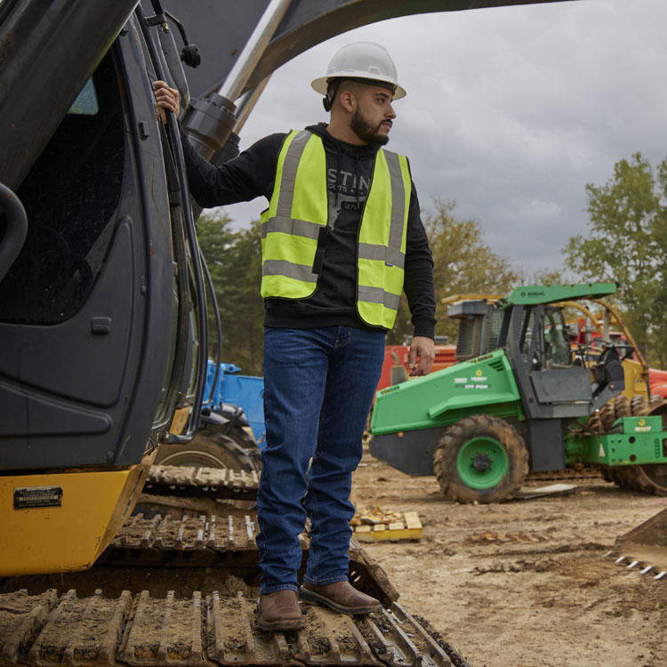 A construction worker wearing a hard hat, safety vest, and jeans stands on heavy equipment at a job site, wearing the Driller 11" work boots.