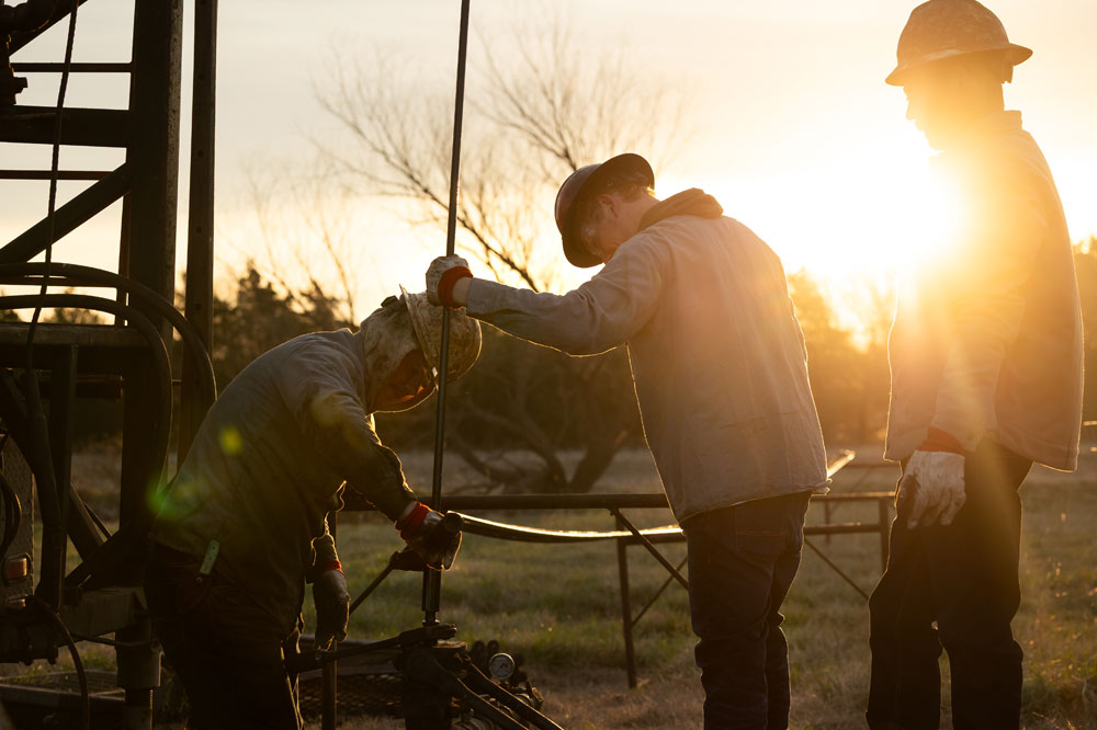 Three men wearing hard hats and covered in oil work on an oil rig, with a sunrise behind them.