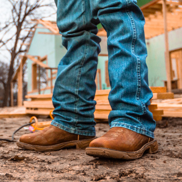 A man wearing the Rush 11” waterproof work boot in saddle tan standing in dirt next to a circular saw