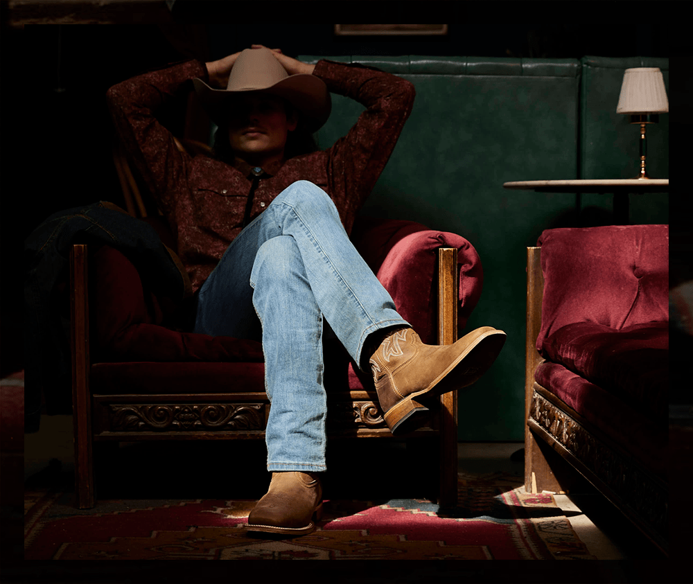 A man with curly hair is wearing a tan cowboy hat, his hands resting above his head, and he is sitting on a maroon couch with brown Frontier Western boots.