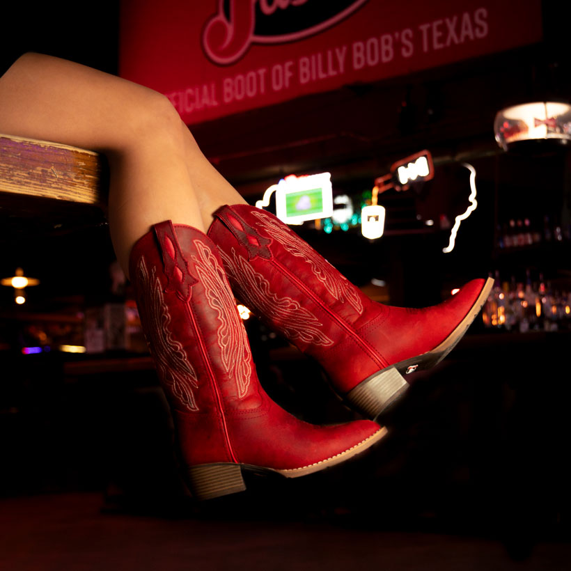 Women’s Renie 12” Western boot in red worn by a woman sitting by a red Justin banner. 