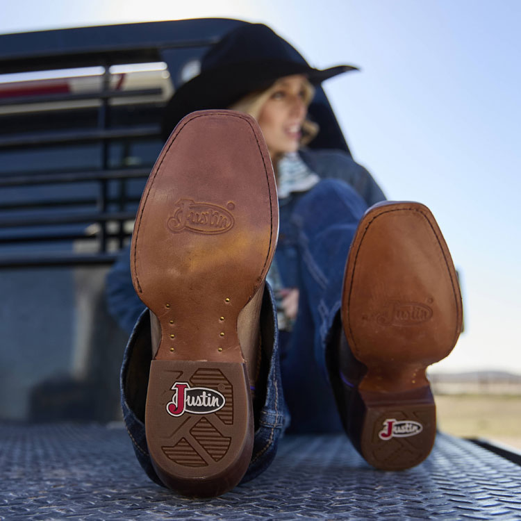 Women’s Annie Up 13” Western boot in tan worn by a blonde woman sitting on the back of a truck. 