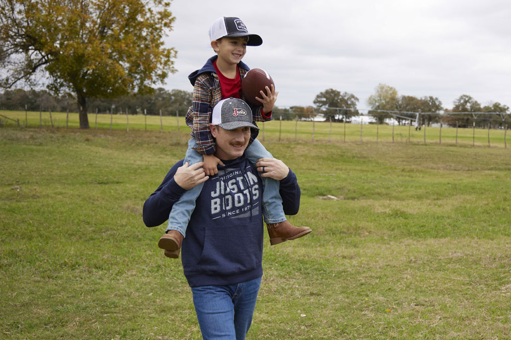 A father carrying his son who is holding a football, sitting on his dad’s shoulders, in a green field, laughing. 