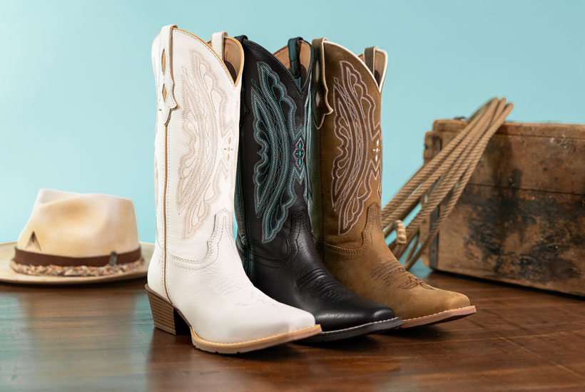 Rae 12” Western boots in brown, Rae 12” Western boots in  black, and Rae 12” Western boots in white, posed in front of a turquoise background and next to a western hat and a rope.
