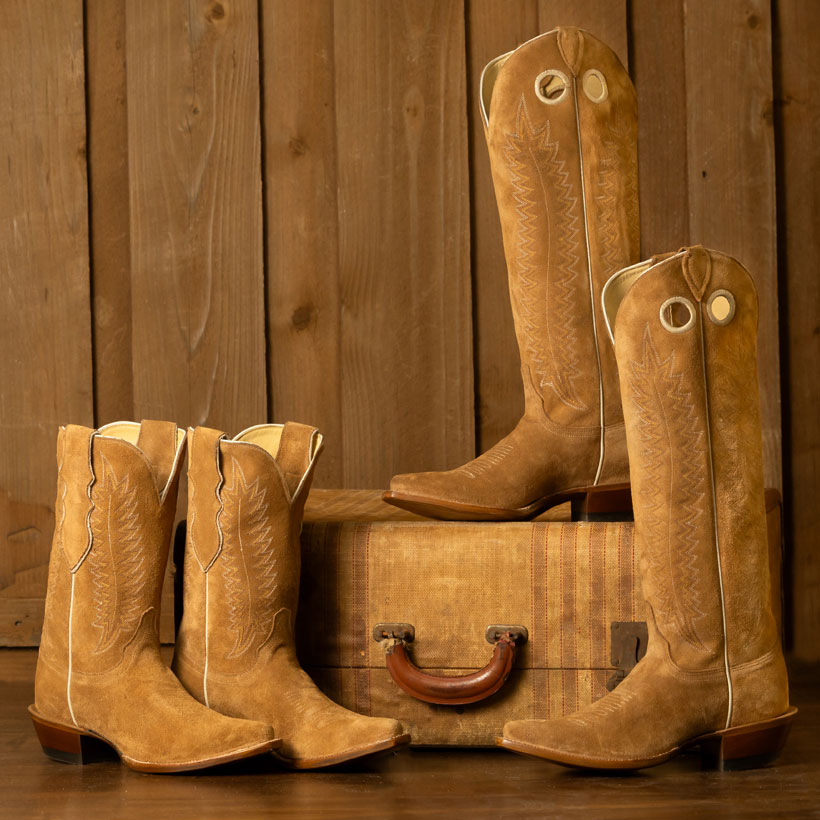 Women’s Sunrise 11” roughout cowgirl boots and Sunset 17” roughout cowgirl boots in tan are posed next to a vintage suitcase. 