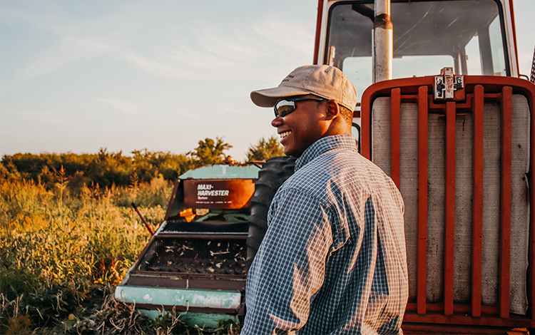 Terren outside in front of tractor wearing a button up shirt, sunglasses and hat.